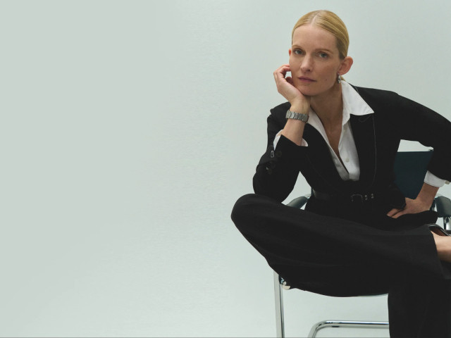 An M.M.LaFleur design: a woman models a sophisticated black pantsuit and white shirt while seated in a chair, conveying confidence and style.